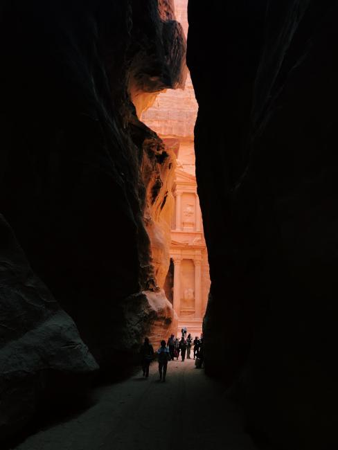 Though the shadows of rocks, the ancient city of petra peeks through in light, with students walking towards it