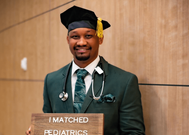 a young Black man wearing doctor's clothes smiles while holding a sign "I matched Pediatrics"
