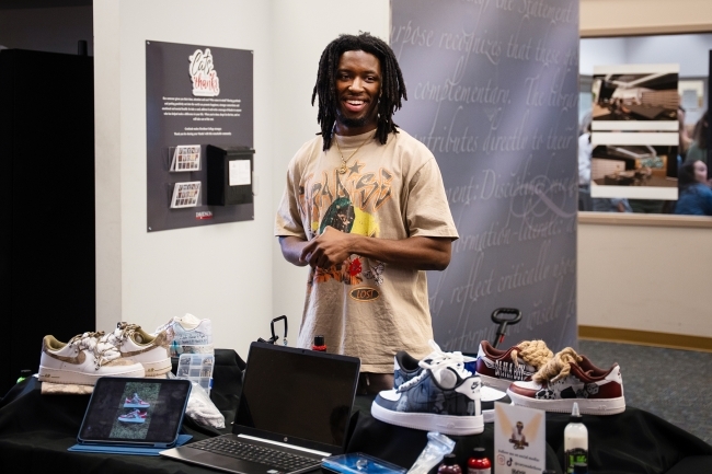 a young man stands behind a table with sneakers and laptops presenting research