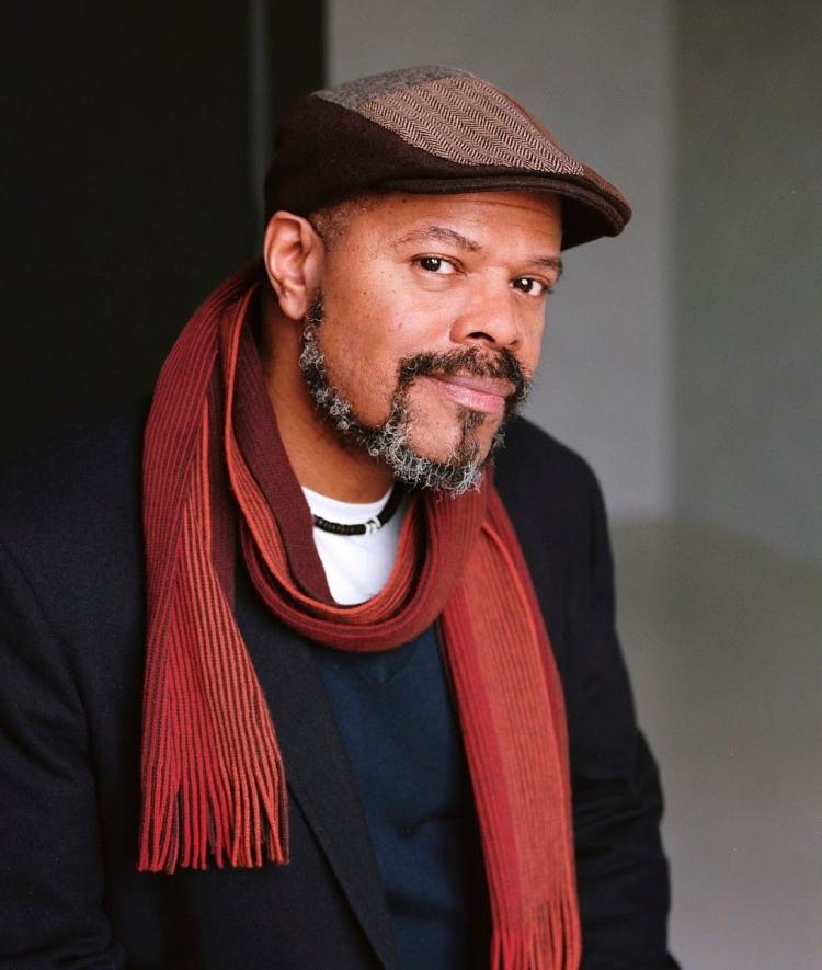 a middle aged Black man wearing a black top and red scarf with a hat while smiling