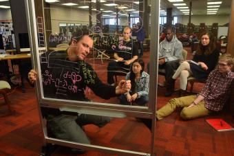Professor Tim Chartier writes on a glass white board while leading a class to a group of students in the library