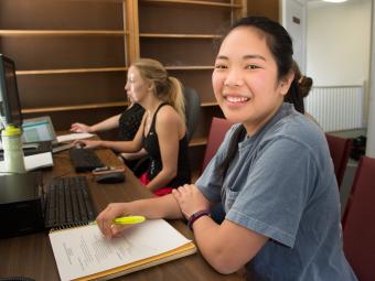 Student smiles at camera in a psychology lab while her peer does research on the computer in the background