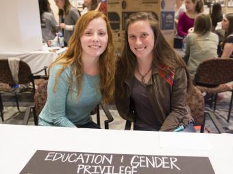 Two students sit at booth with a poster focused at education and gender privilege