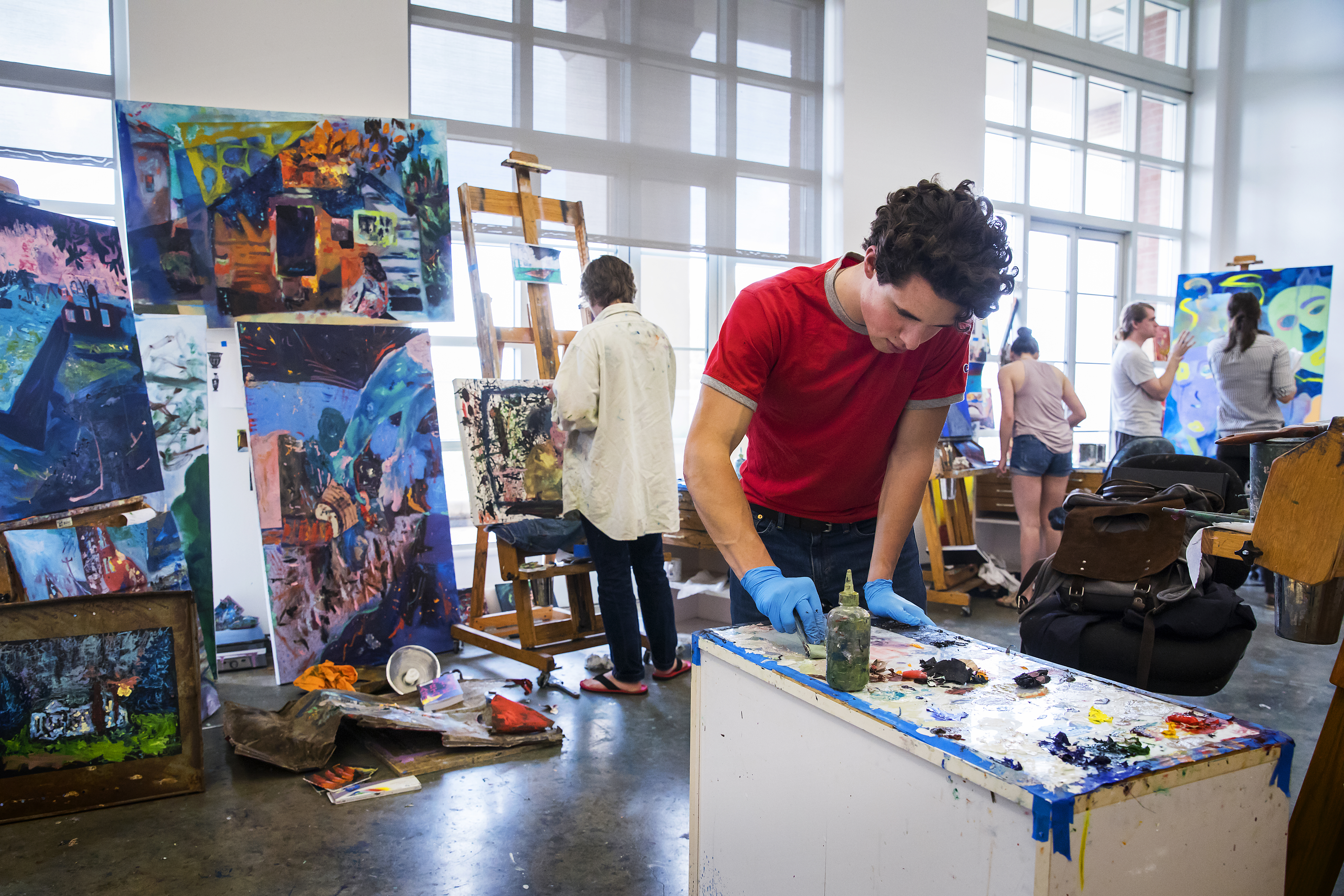 Student mixes paint in foreground while other students paint canvases in background