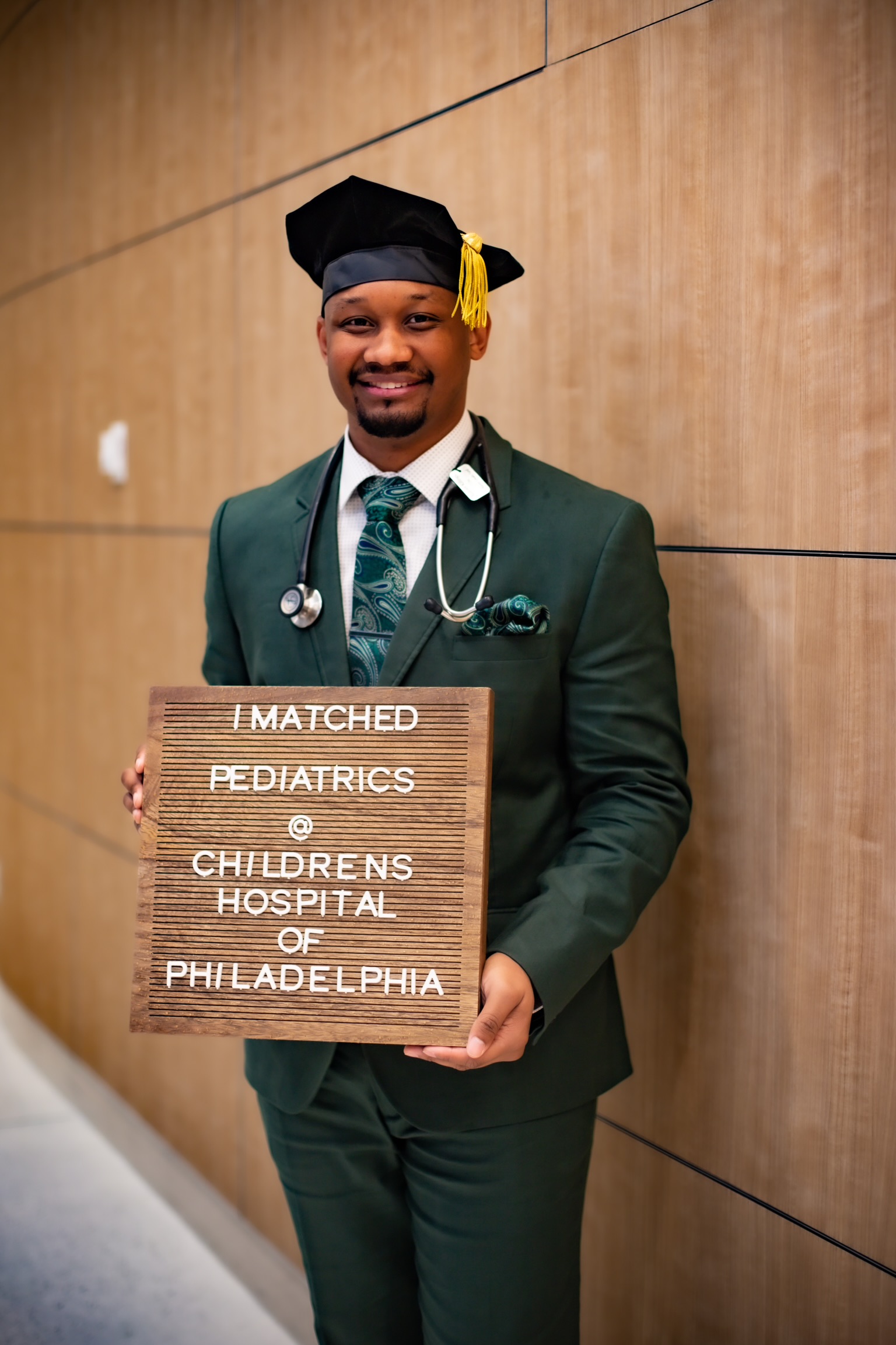 a young man wears a graduation cap and suit and holds a sign that says "I matched Pediatrics at Childrens Hospital of Philadelphia"