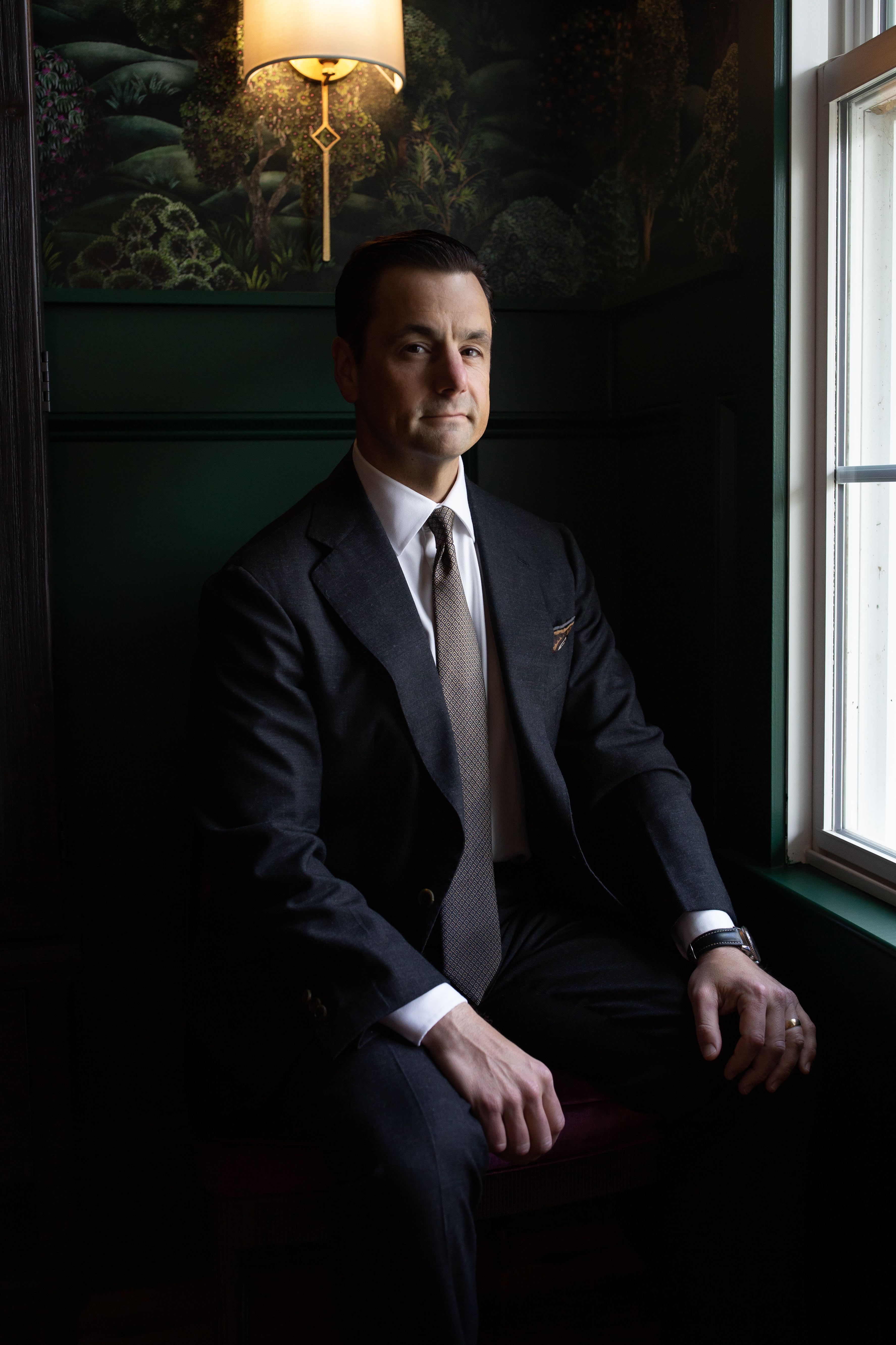 a young white man wearing a suit sitting in a dark room with a lamp behind