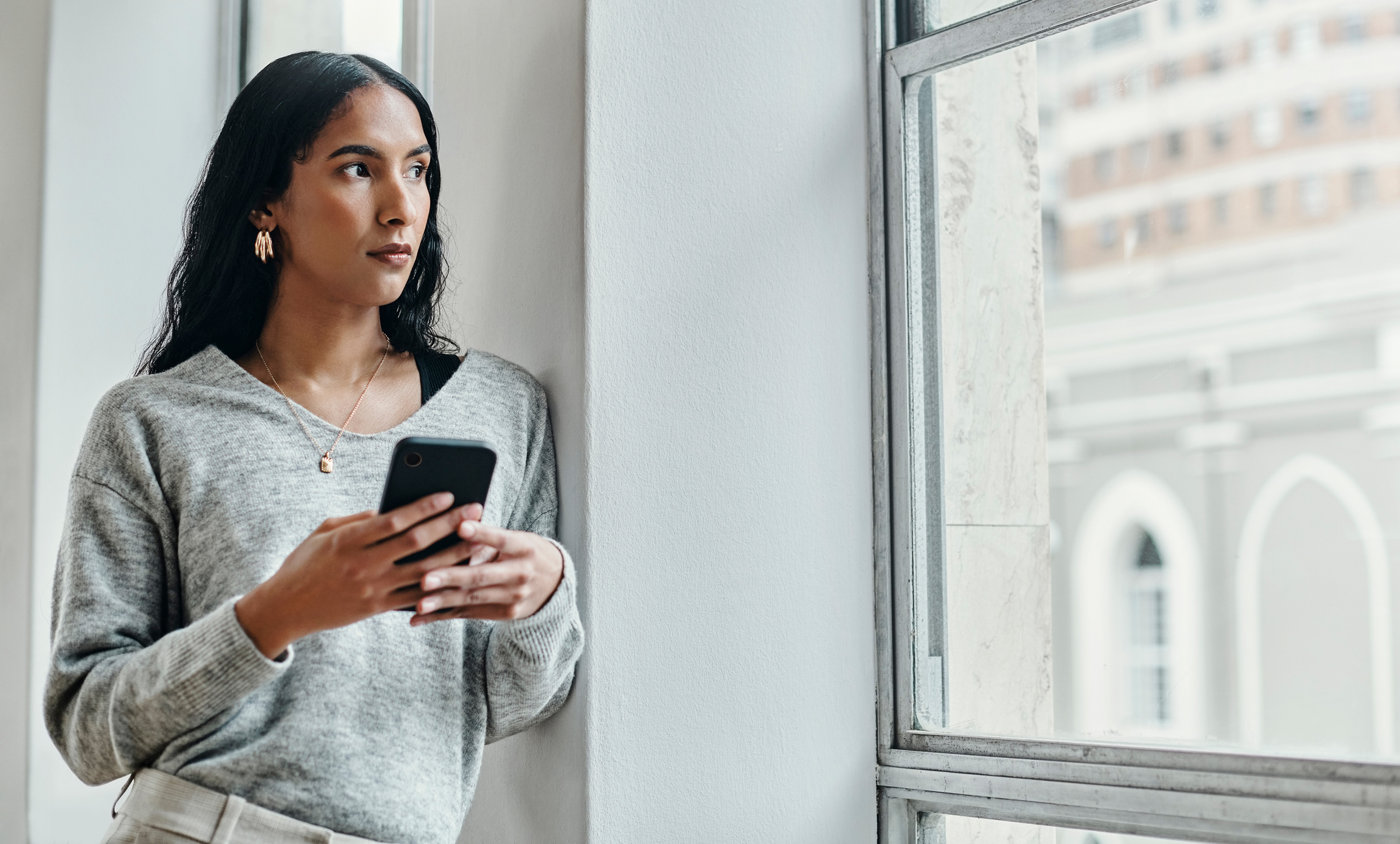 Woman holding a cell phone looking out the window wondering about social media misinformation