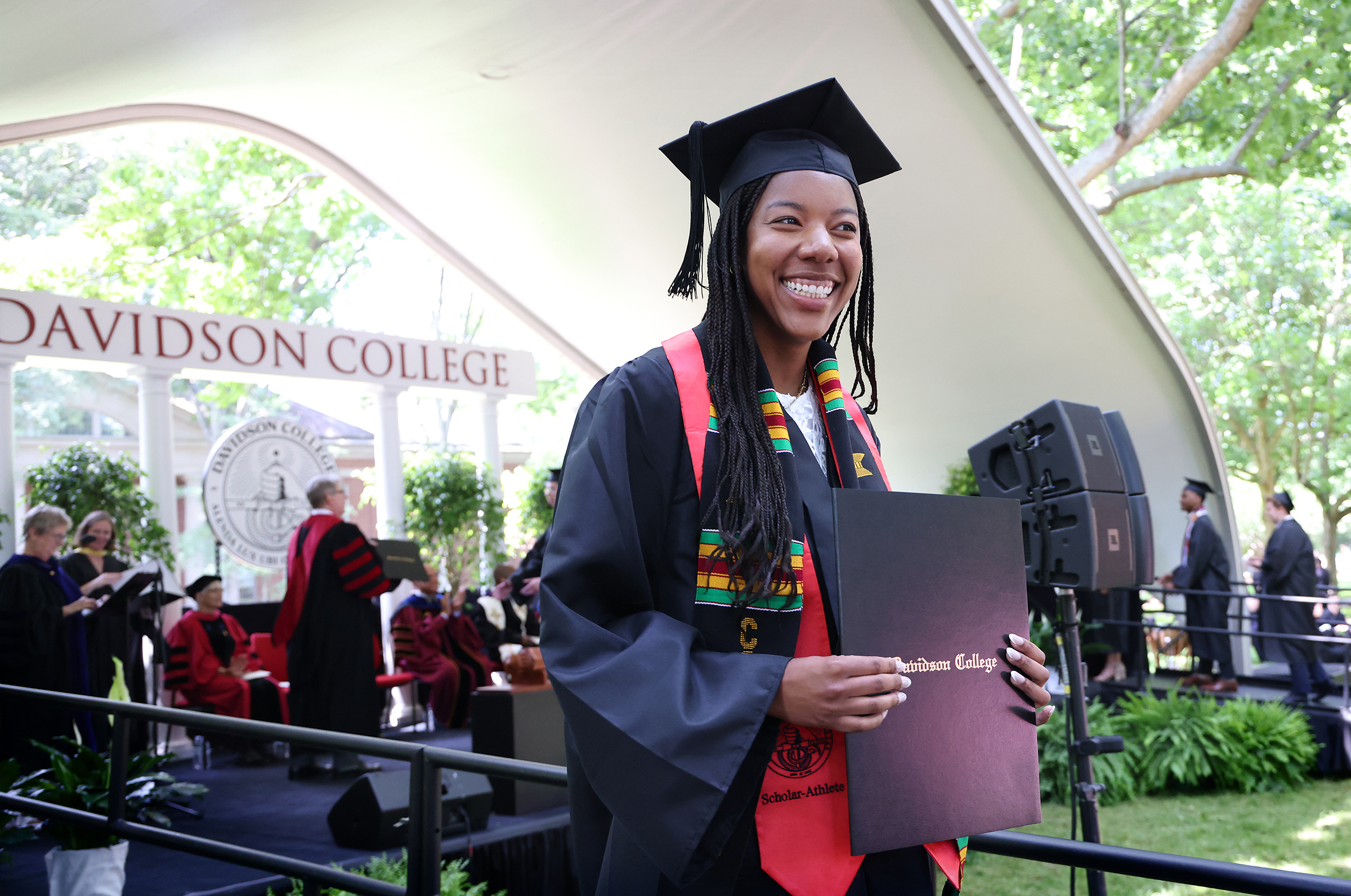 a young Black woman holds a diploma while wearing graduation robes