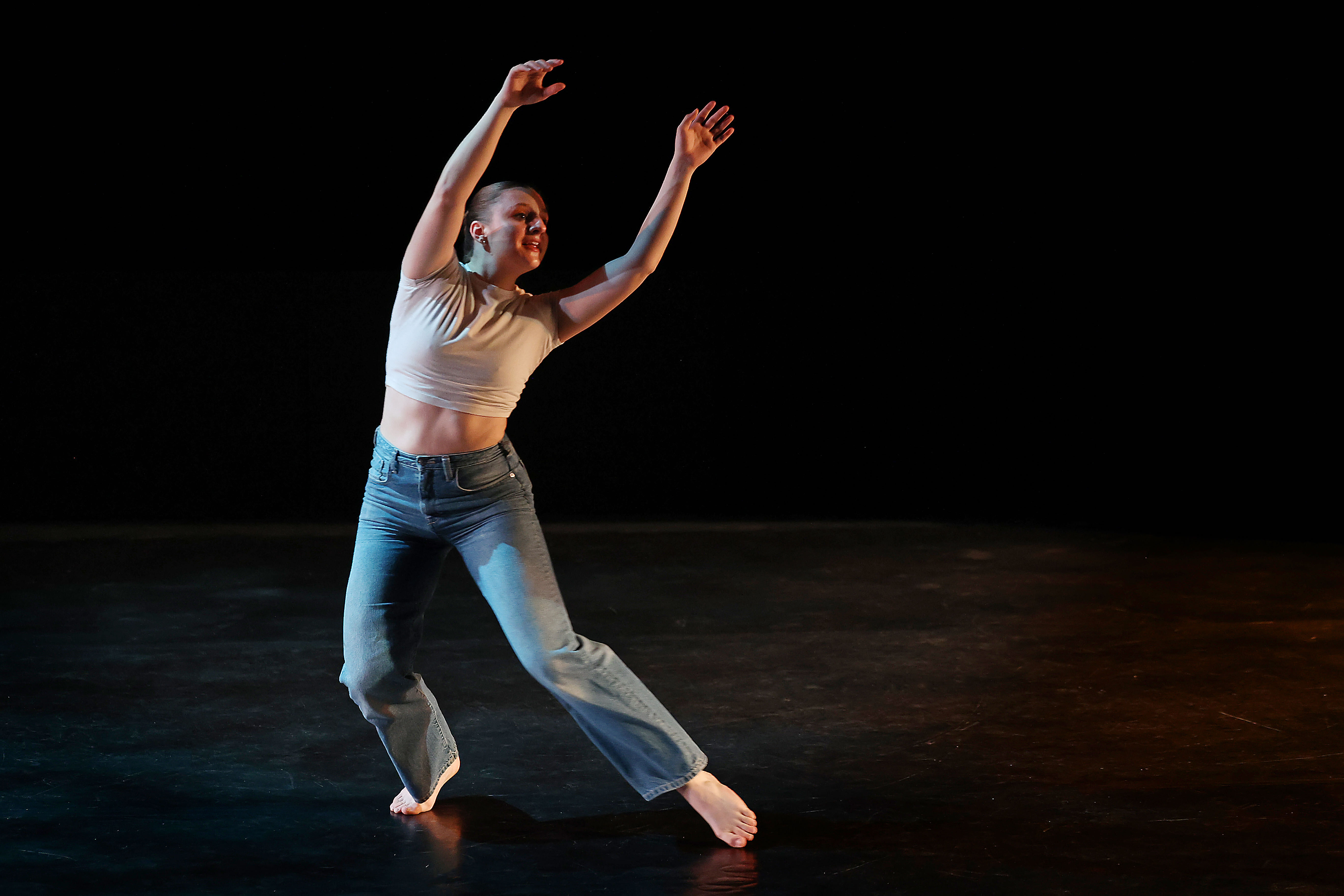 a young woman dances in a white top and jeans on a black stage