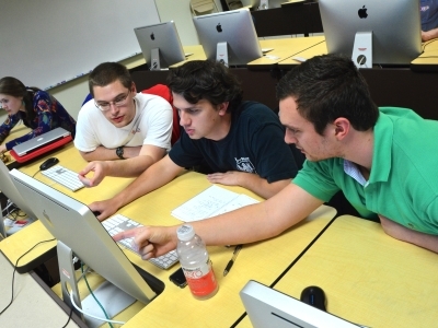 Three students in genomics class sit in computer lab and point at a desktop screen while discussing