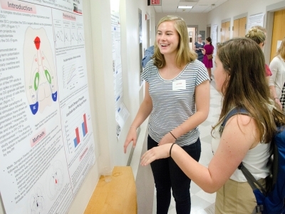 Student presents on her research standing by a poster and talking to a fellow student