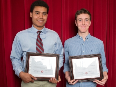 Two students stand in front of red curtain holding their economics awards