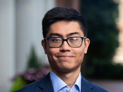 A young man wearing a jacket and collared shirt and glasses smiles