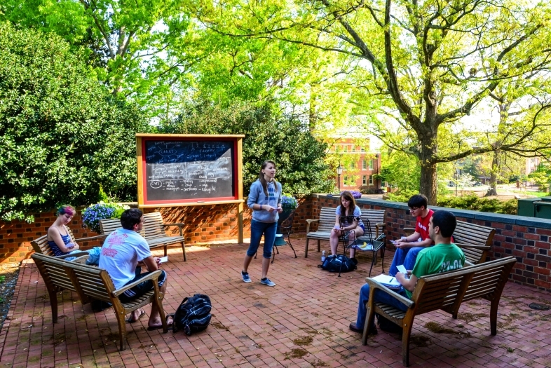 Russian Studies AT Session in outdoor classroom where students sit on benches and instructor addresses them in front a chalkboard with writing in Russian