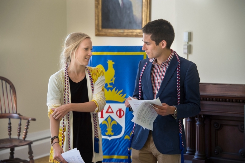 Two students stand in front of a colorful flag for Pi Delta Phi talking with honors cords around their necks