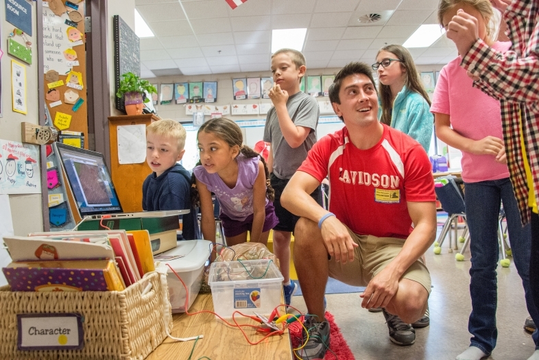 Davidson student wearing a Davidson soccer jersey stands amidst a group of young kids in their classroom and shows them math exercises on a laptop