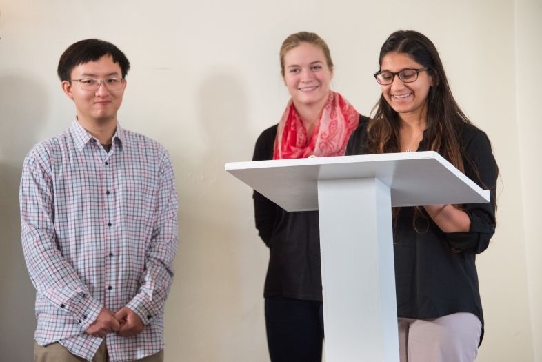 Two students stand by a podium addressing the audience while another stands off to the side