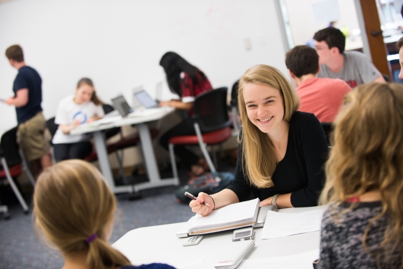 Student talks to classmates sitting at same table. Rest of class is out of focus in the background