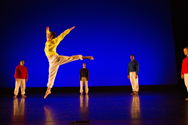 Student leaps into the air in front of blue lights while other dancers on stage stand still