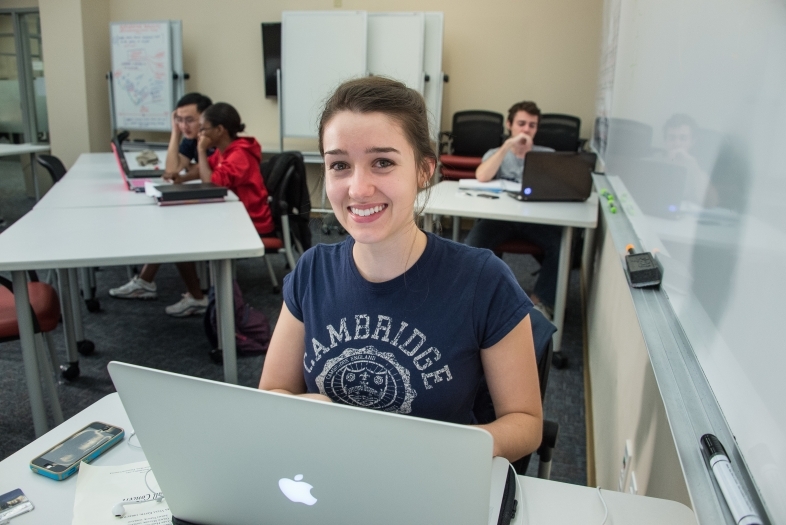 Student smiles at camera with tutoring supplies in front of her and people being tutored in the background