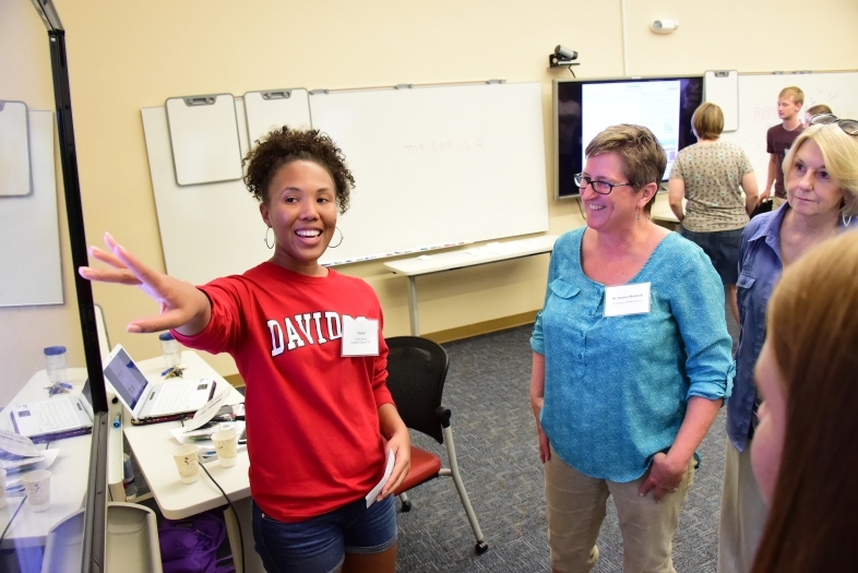 Student stands near screen and points while crowd gathers around her and hears her share