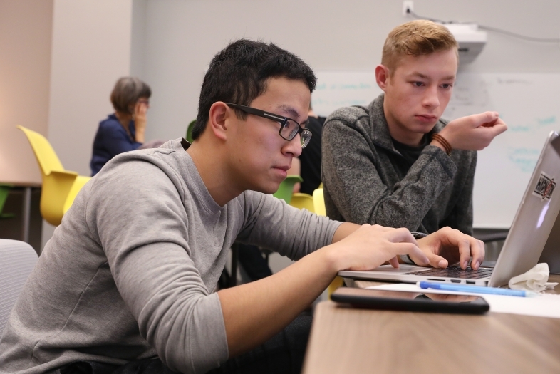 Students sit at table and while one types on keyboard, other student watches