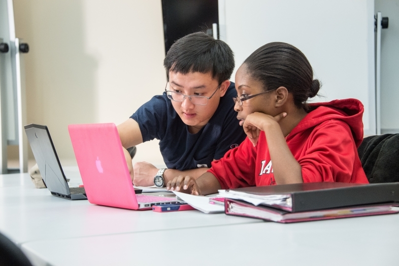 Two students talk with laptops open, one pointing at his colleague's laptop