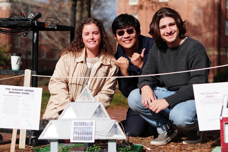 three students smiling with a tiny house in front of them