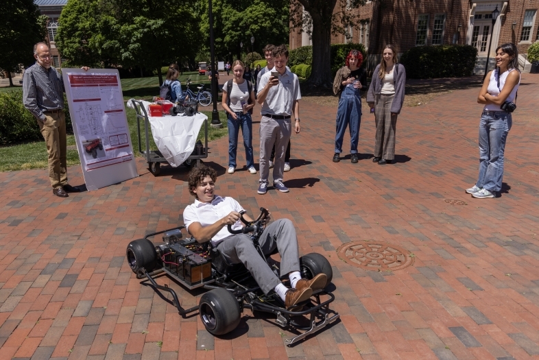 A group of students and faculty stand around and smile as a student drives a go-kart