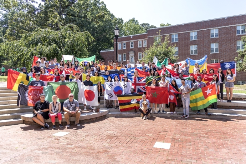 A large group of students in front of a brick building holding flags from around the world