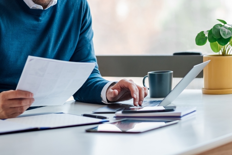 a person sits at a white desk with a laptop and stack of paper