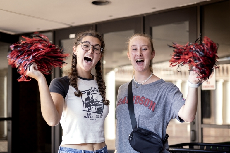 two young women hold pom poms and smile while cheering