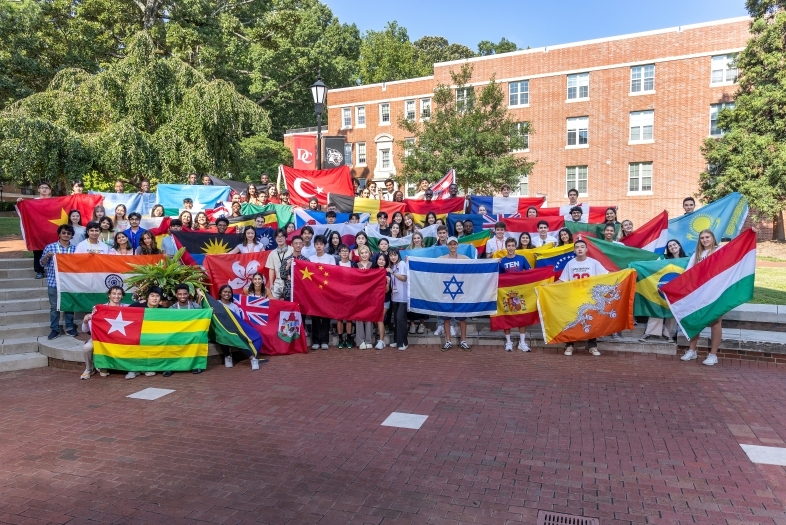 a group of students holding flags from around the world outside a brick college building