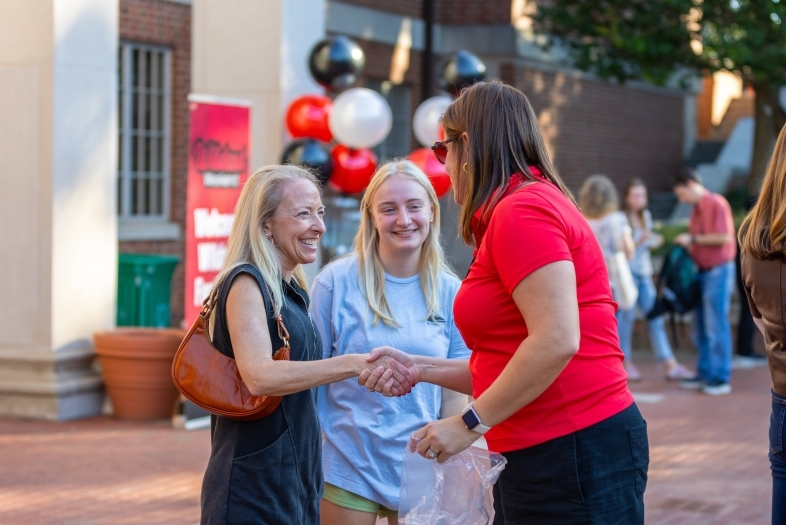 three women greet each other on a brick sidewalk in front of an academic building