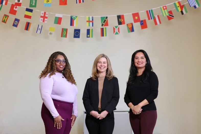 a group of three women stand together in front of a garland of international flags