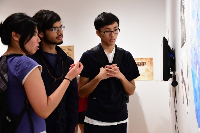 Three students huddle around art work displayed in the gallery and discuss