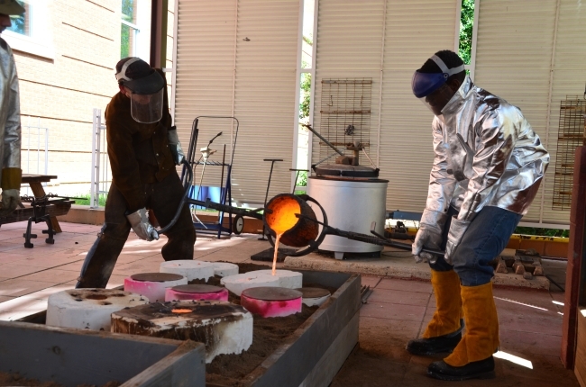 Professor and student dressed in protective gear pour liquid bronze at the bronze foundry.