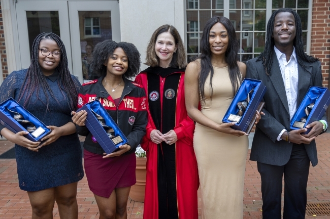 Four students holding their trophy awards next to President Carol Quillen in the union amphitheatre