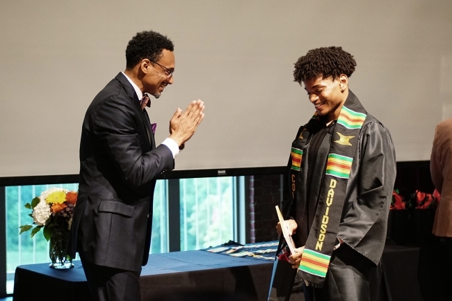 Two Black men smile as one puts graduation regalia around himself