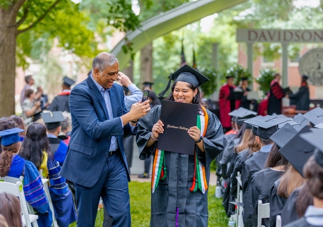 Student holds diploma and smiles while father takes picture