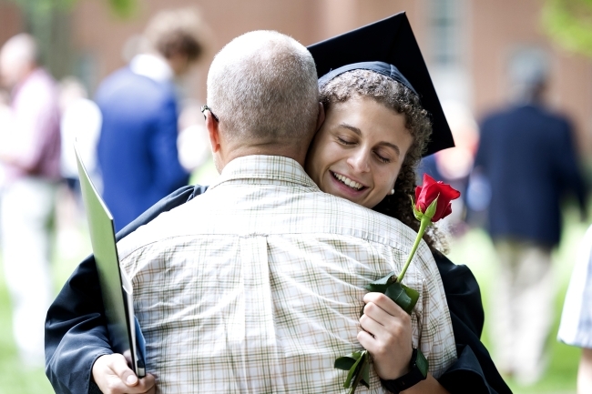 Female student in cap and gown embraces her father while holding a flower