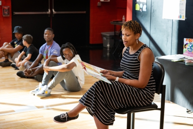 a staff member reads a book to students sitting in a gym