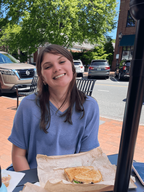 a young woman with brown hair sits at a table on a main street with a sandwich in front of her