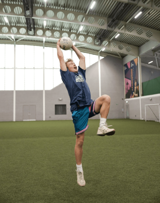 a male wearing athletics throws a soccer ball while in an indoor field