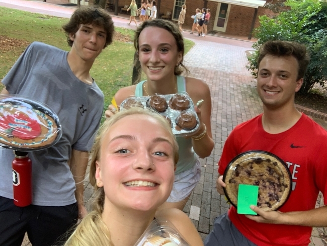 a group of four students hold up cakes while standing outside