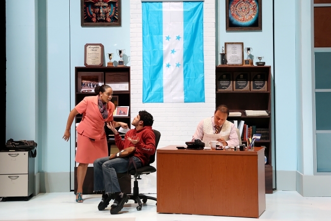 on stage, a woman in a suit stands over and yells at a student while a male teacher sits by at a desk