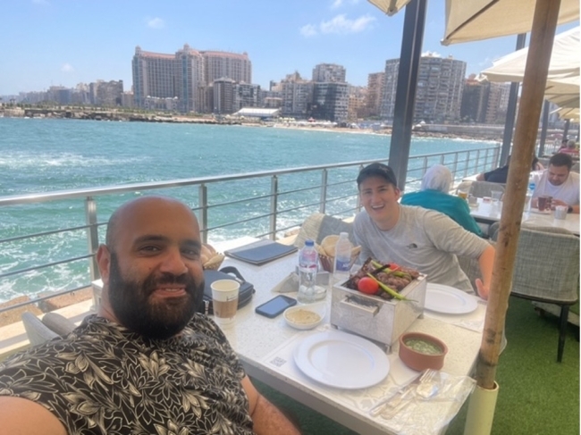 a young man and an older man sit at a dining table overlooking the ocean