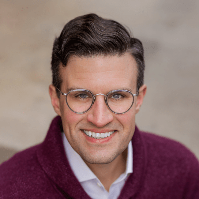 a young white man with glasses wearing a purple sweater and smiling