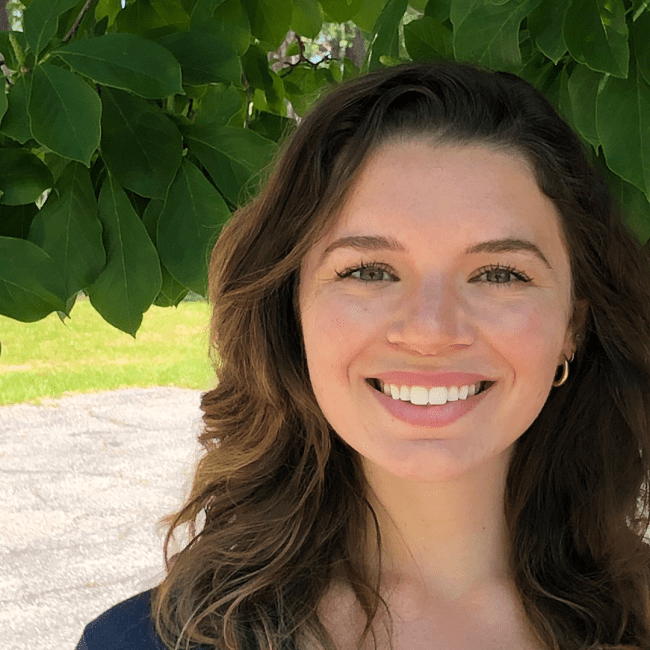 a young woman with brown hair smiles with a leafy tree in the background