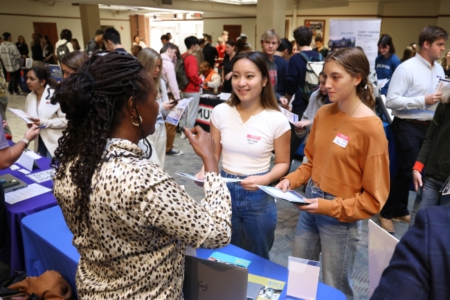 two young women talk to an older woman while holding papers at a job fair